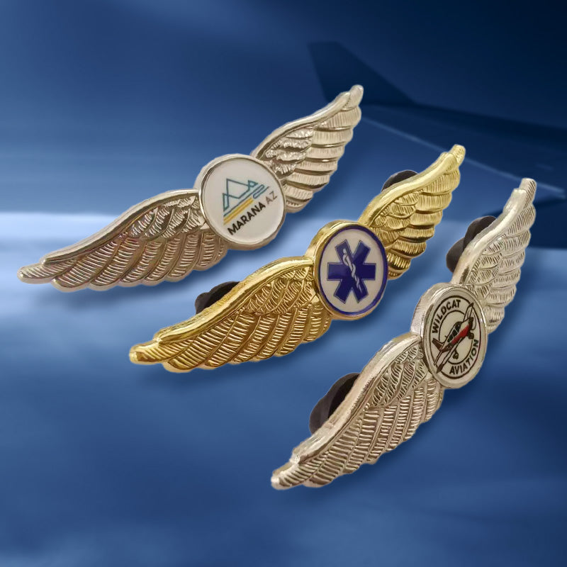Three pilot wings lapel pins in side view, featuring mixed gold and silver-tone finishes with distinct circular emblems: one displaying the "MARANA A" logo, another with a purple medical caduceus symbol, and the third showcasing "BLUECAT AVIATION" with a fish graphic — arranged against a blue sky background.