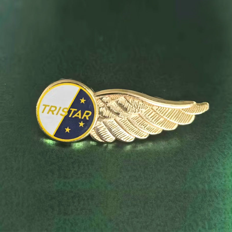 Gold-tone pilot wings lapel pin in side view, featuring a circular emblem divided into white and navy blue enamel sections with "TRISTAR" text and three gold stars, displayed against a dark green background.