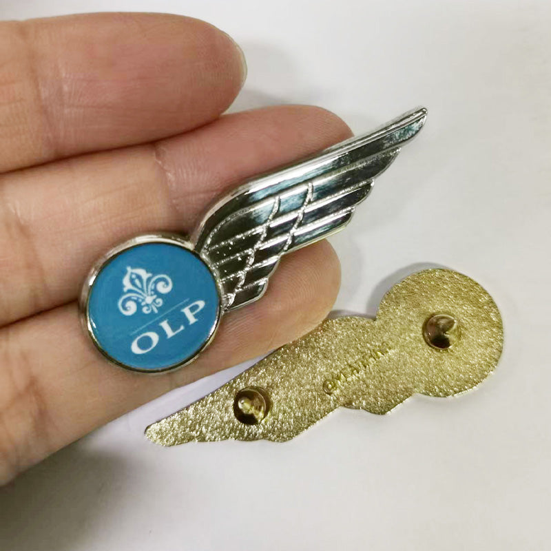 Hand holding a silver-tone pilot wings lapel pin in side view, featuring a blue circular emblem with a white fleur-de-lis symbol and the text “OLP,” displayed above its gold-tone backside with visible pin-back hardware and branding.
