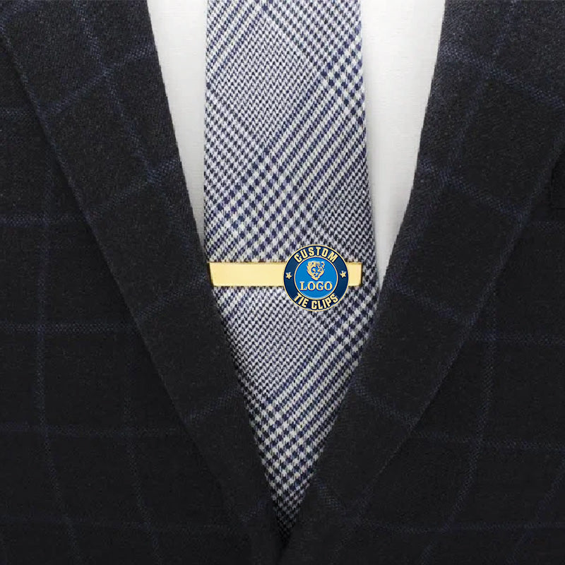 Custom gold tie clip with blue enamel emblem, featuring custom text, shown on a herringbone pattern tie in a professional business attire scene.