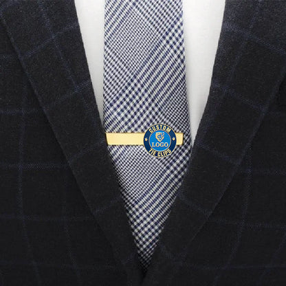 Custom gold tie clip with blue enamel emblem, featuring custom text, shown on a herringbone pattern tie in a professional business attire scene.