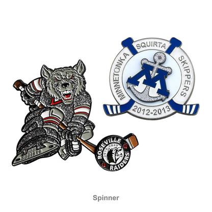 Two custom soft enamel spinner hockey trading pins on a white background: left pin features a silver glitter wolf player with a hockey stick, right pin is a blue and white circular design with an anchor and letter M, both with spinning elements and custom team text.