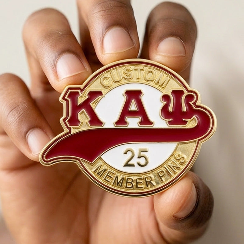 A hand holding a circular gold KAP pin featuring the Greek letters Kappa Alpha Psi in crimson and white with the text Custom 25 Member Pins.
