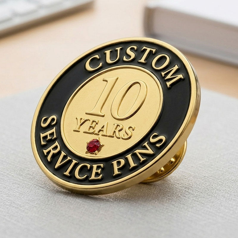 A side-angle perspective view of a custom round 10-year service recognition lapel pin in gold tone, resting on a textured surface, showing its polished golden center with a red gemstone accent and a black enamel border with raised lettering.