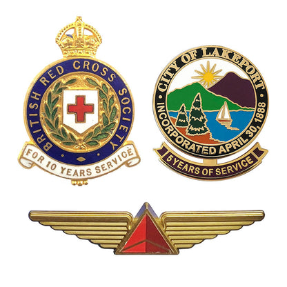 A display of three custom milestone pins, featuring a crown-topped British Red Cross Society 10 years service badge, a colorful round City of Lakeport 5 years of service pin with a sailboat scene, and a gold-tone aviator wing pin with a central red triangle logo.