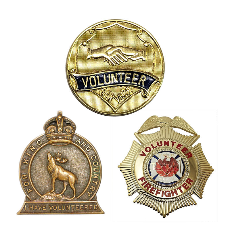 A display of three custom volunteer service pins, showcasing a round textured gold-tone pin with a handshaking design, an antique bronze military-style badge featuring a crown and a stag, and a starburst-shaped gold-tone volunteer firefighter shield with red enamel text.