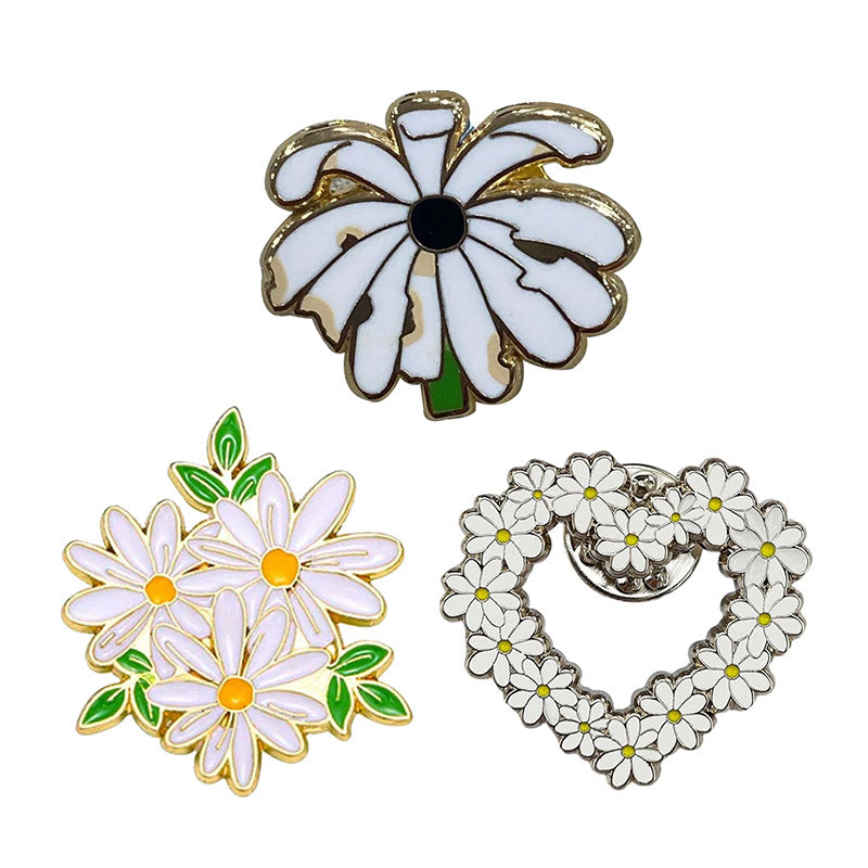 A trio of custom daisy lapel pins, featuring a single white daisy bloom pin with a black center, a gold-tone bunch of light purple daisies with green leaves, and a heart-shaped wreath pin made entirely of tiny white and yellow daisies.