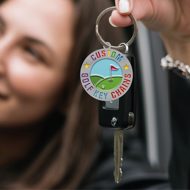Custom golf keychain with a circular metal badge featuring custom text, held in a hand with white nail polish attached to black car keys against a blurred car interior background
