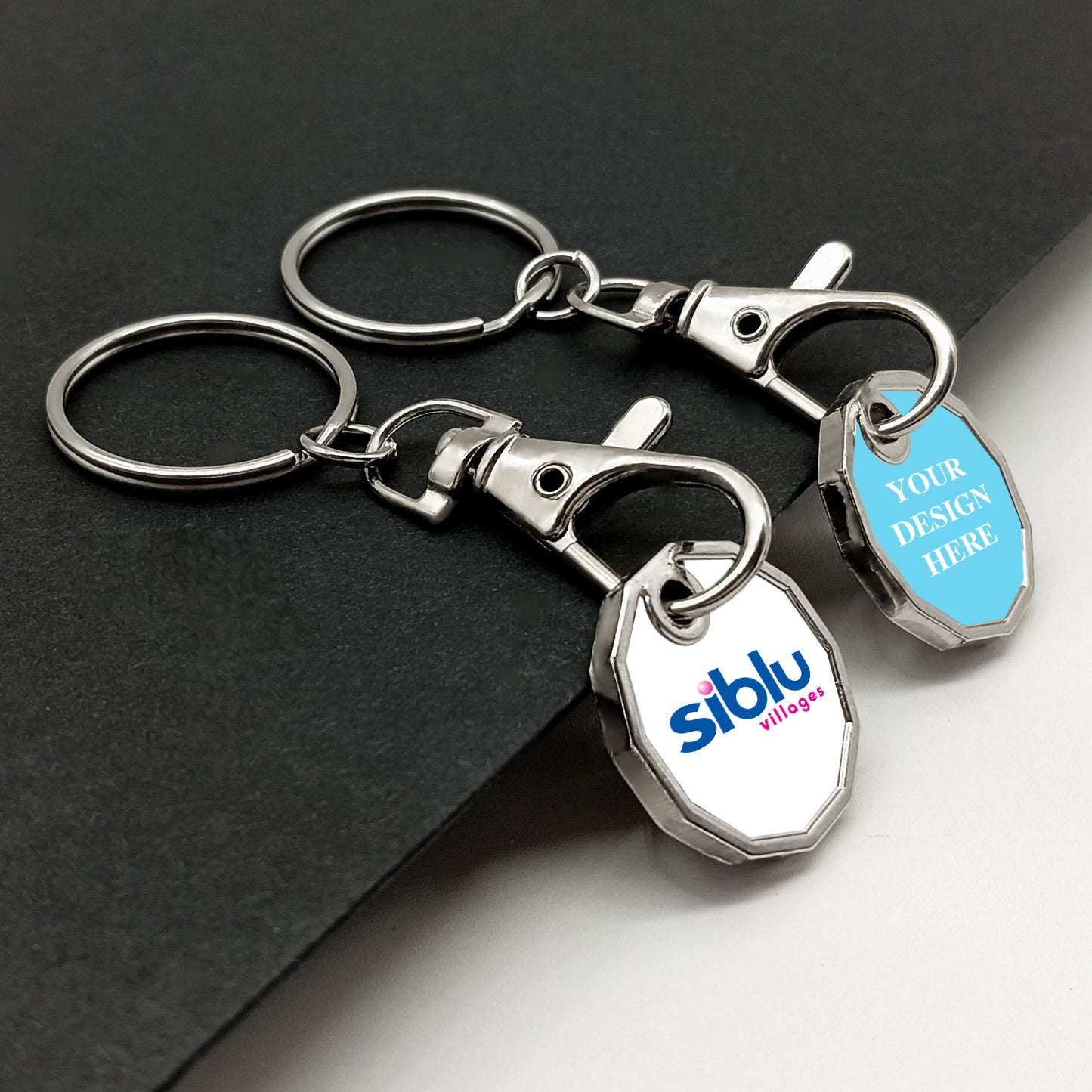 A pair of metal trolley token keyrings with chrome finish, placed side by side. The left tag shows a logo, and the right tag displays placeholder text on a light blue background, demonstrating personalization potential. They rest on a dark textured surface.