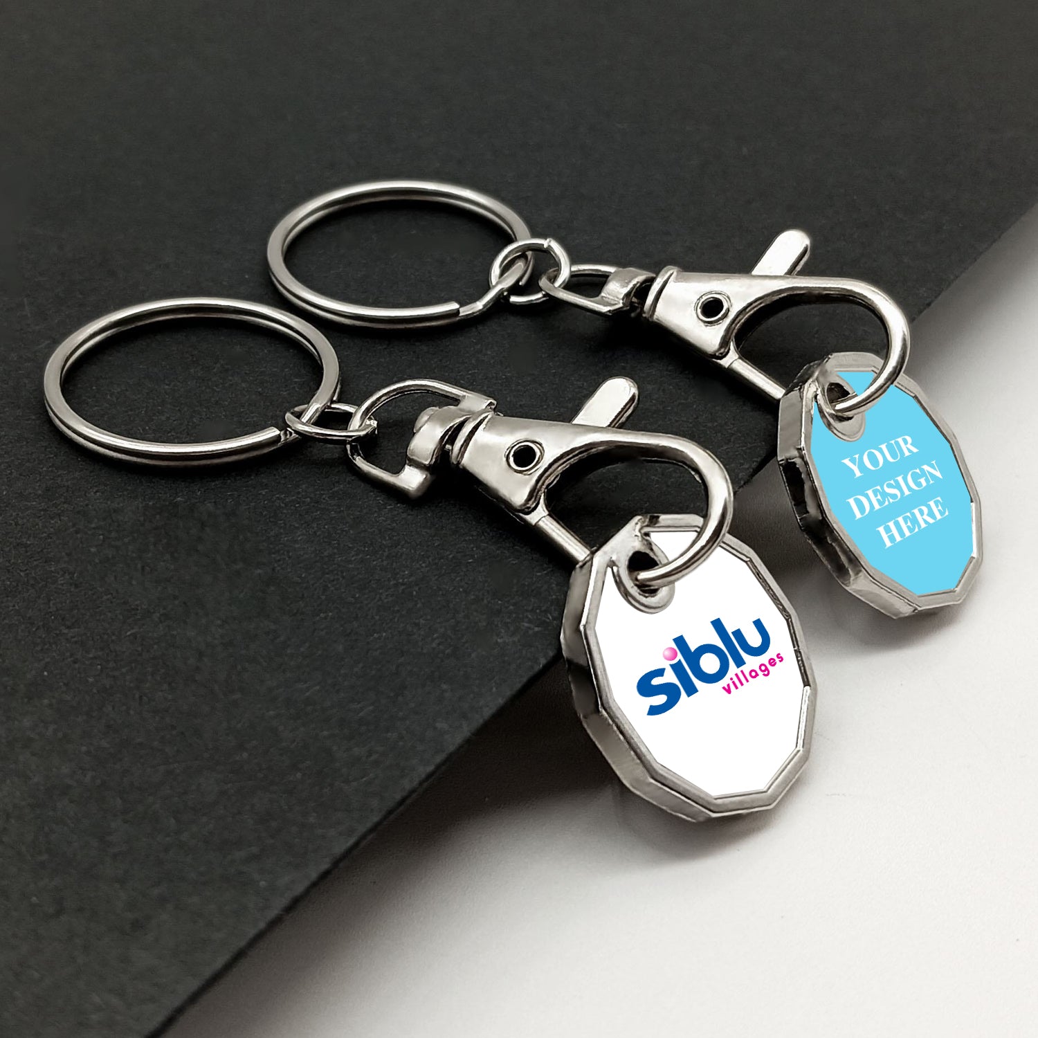 A pair of metal trolley token keyrings with chrome finish, placed side by side. The left tag shows a logo, and the right tag displays placeholder text on a light blue background, demonstrating personalization potential. They rest on a dark textured surface.
