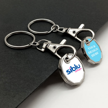 A pair of metal trolley token keyrings with chrome finish, placed side by side. The left tag shows a logo, and the right tag displays placeholder text on a light blue background, demonstrating personalization potential. They rest on a dark textured surface.