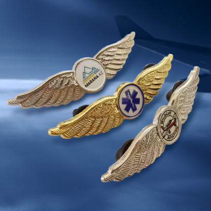 Three pilot wings lapel pins in side view, featuring mixed gold and silver-tone finishes with distinct circular emblems: one displaying the "MARANA A" logo, another with a purple medical caduceus symbol, and the third showcasing "BLUECAT AVIATION" with a fish graphic — arranged against a blue sky background.