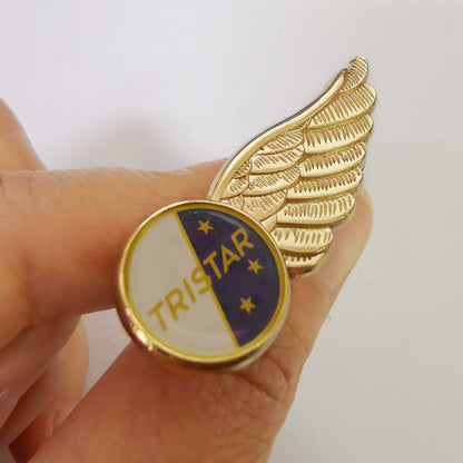 Hand holding a gold-tone pilot wings lapel pin in side view, featuring a circular center divided into navy blue and white enamel sections with “TRISTAR” text and three stars — showcasing detailed feather textures and classic aviation-inspired design.