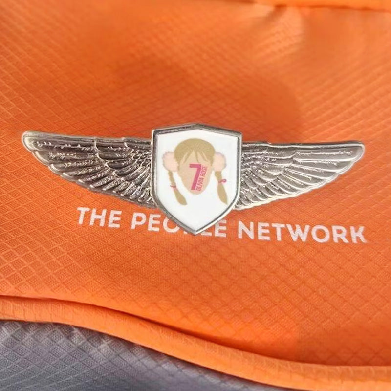 Silver-tone pilot wings lapel pin featuring a central shield emblem with a cartoon-style illustration of a girl with pigtails, a large number “7”, and the word “SEVEN” beneath it, worn on an orange textured fabric uniform above the text “THE PEOPLE NETWORK”.