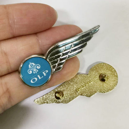 Hand holding a silver-tone pilot wings lapel pin in side view, featuring a blue circular emblem with a white fleur-de-lis symbol and the text “OLP,” displayed above its gold-tone backside with visible pin-back hardware and branding.