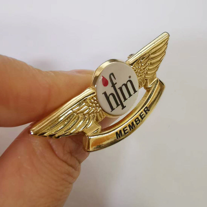 Hand holding a gold-tone pilot wings lapel pin in side view, featuring a circular center with “hfm” text and a red droplet accent, plus a curved banner below engraved with “MEMBER” — showcasing detailed feather textures and premium metallic finish.