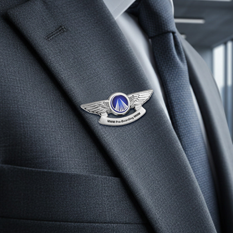 Silver-tone pilot wings lapel pin worn on the lapel of a dark gray suit jacket, featuring a blue circular emblem with a white triangular runway design and a silver banner below inscribed with “MWM Pre-Boarding 1988” — displayed against a softly blurred indoor background with natural light.