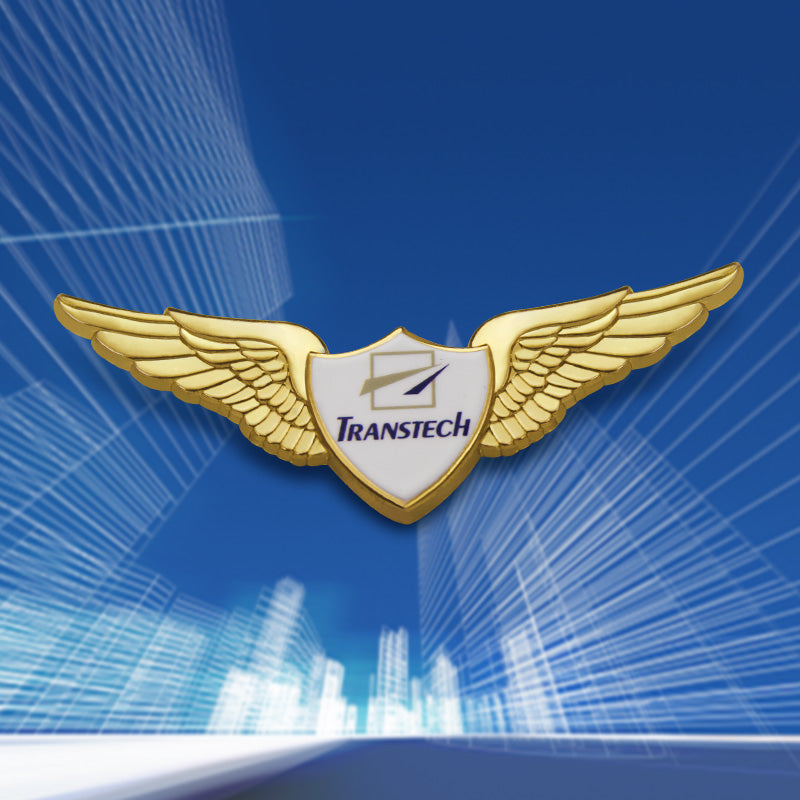 Gold-tone pilot wings lapel pin displayed against a dynamic blue background with abstract cityscape lines, featuring a central white shield emblem with the “TRANSTECH” logo in purple and a stylized lightning bolt — showcasing polished metallic wings with finely detailed feather textures in a front-facing perspective.