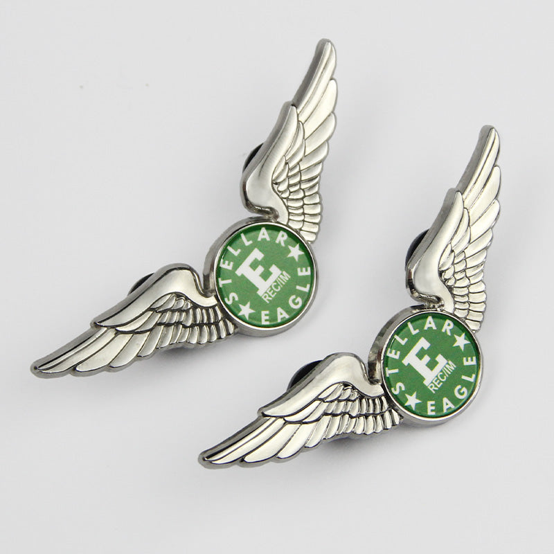 Hand holding two silver-tone pilot wings lapel pins in side view, each featuring a green circular emblem with "STELLAR EAGLE REGIM" and a bold "E" center — showcasing detailed feather textures and pin-back closures for secure attachment.