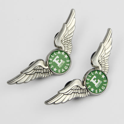Hand holding two silver-tone pilot wings lapel pins in side view, each featuring a green circular emblem with "STELLAR EAGLE REGIM" and a bold "E" center — showcasing detailed feather textures and pin-back closures for secure attachment.