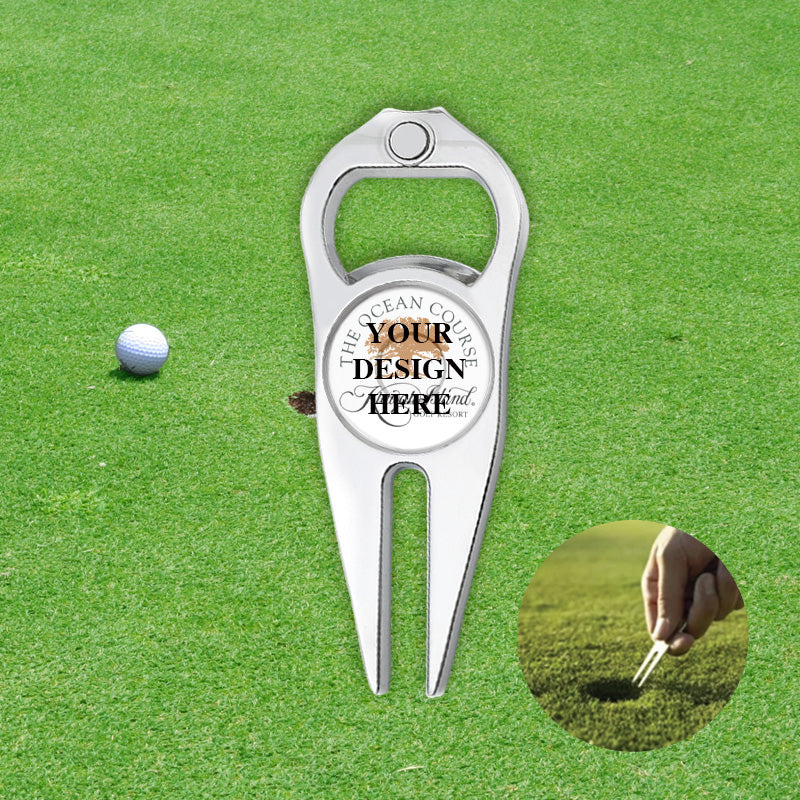 On the green of a golf course, with a white golf ball on the left and a close-up of a hand holding a Personalized Golf Divot Tool in the lower right corner.