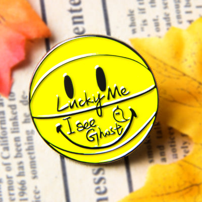 A custom metal pin with a yellow background featuring a smiley face and the text 'Lucky Me I see Ghost', placed on a surface with autumn leaves and a patterned background.
